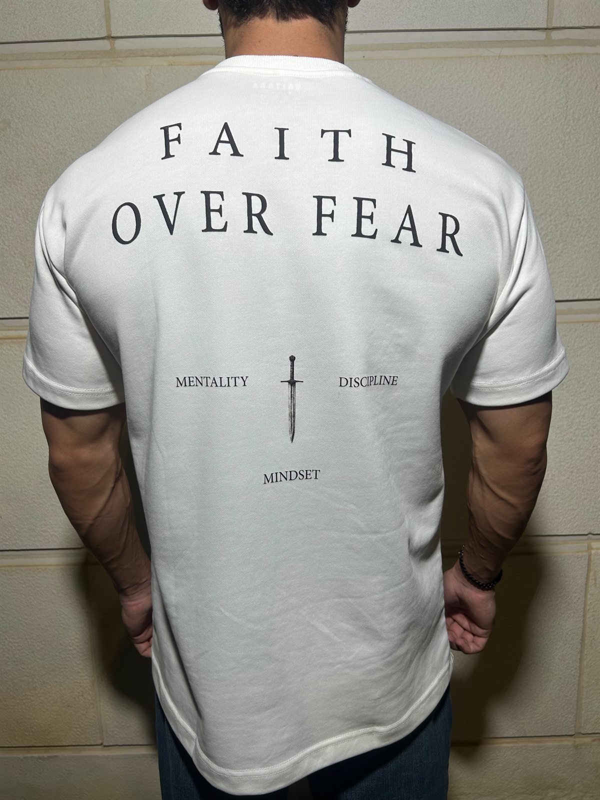 The back of the white Faith Over Fear tee, FAITH OVER FEAR over a dagger with mentality, discipline and mindset around it